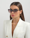 A person with long brown hair wearing a white blazer and 18-karat gold-plated jewelry, including a chain necklace and hoop earrings, is pictured against a light gray background. They are donning large Gaia - Champagne sunglasses by For Art's Sake®, which are handmade from plant-based acetate and have a serious expression on their face.