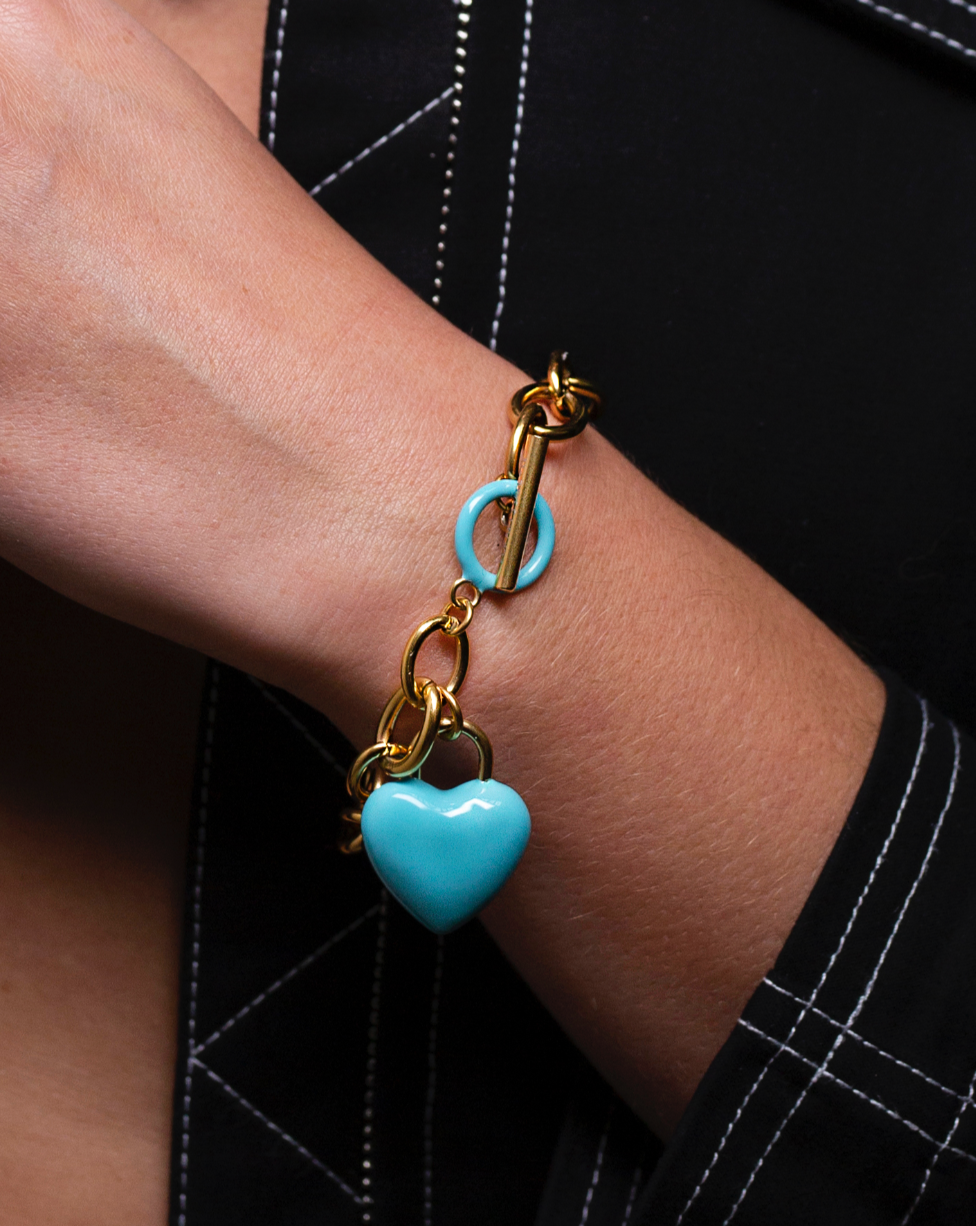 A close-up of a person's wrist adorned with The Kiss Bracelet Blue by For Art's Sake®, featuring a chunky 18k gold bracelet with a turquoise circular charm and a dangling enamel heart charm. The person is wearing a dark outfit with white stitching.