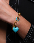 A close-up of a person's wrist adorned with The Kiss Bracelet Blue by For Art's Sake®, featuring a chunky 18k gold bracelet with a turquoise circular charm and a dangling enamel heart charm. The person is wearing a dark outfit with white stitching.