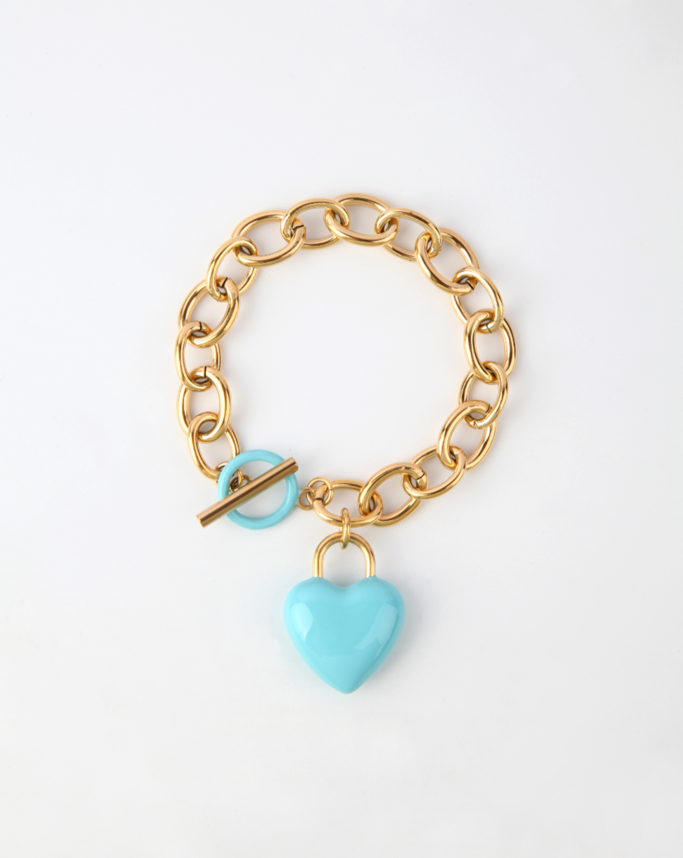 A gold chain bracelet with a large link design features an 18k gold, turquoise enamel heart-shaped charm. The bracelet has a toggle clasp, with the toggle bar passing through a matching turquoise loop. "The Kiss Bracelet Blue" by For Art's Sake® is displayed on a plain white background.