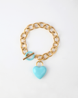 A gold chain bracelet with a large link design features an 18k gold, turquoise enamel heart-shaped charm. The bracelet has a toggle clasp, with the toggle bar passing through a matching turquoise loop. "The Kiss Bracelet Blue" by For Art's Sake® is displayed on a plain white background.