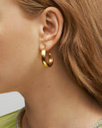 Close-up of a person's ear adorned with Wave Earrings Silver from For Art's Sake®. The person's light brown hair is tucked behind the ear, revealing the earring fully. Part of a light green top with a sparkly neckline can be seen. The background is a neutral gray.