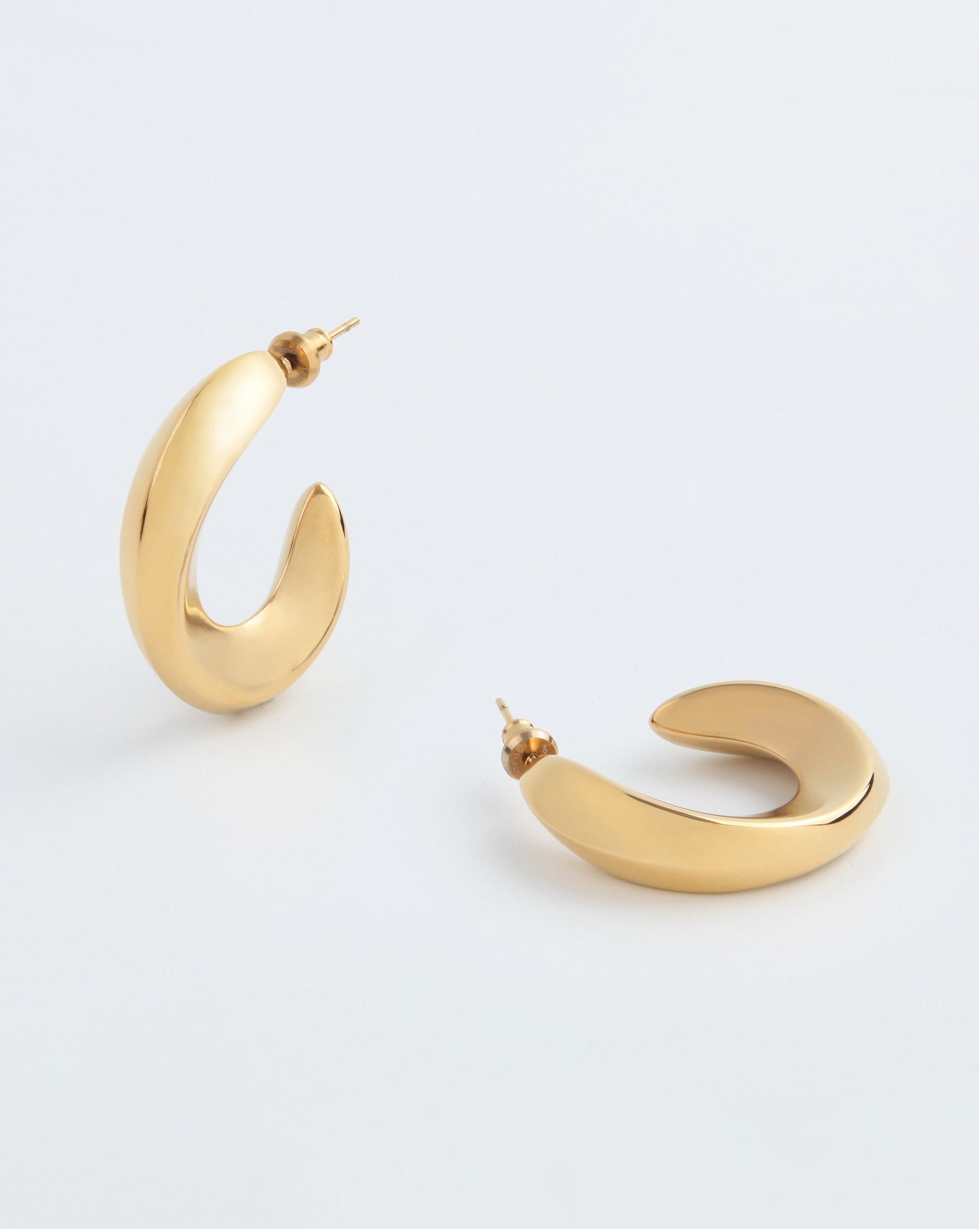 A pair of crescent-shaped, chunky half hoop design gold Wave Earrings Gold by For Art's Sake® is shown against a plain white background. One 18kt gold-plated earring is standing upright, and the other is laid flat, showcasing their smooth and hand-polished finish.