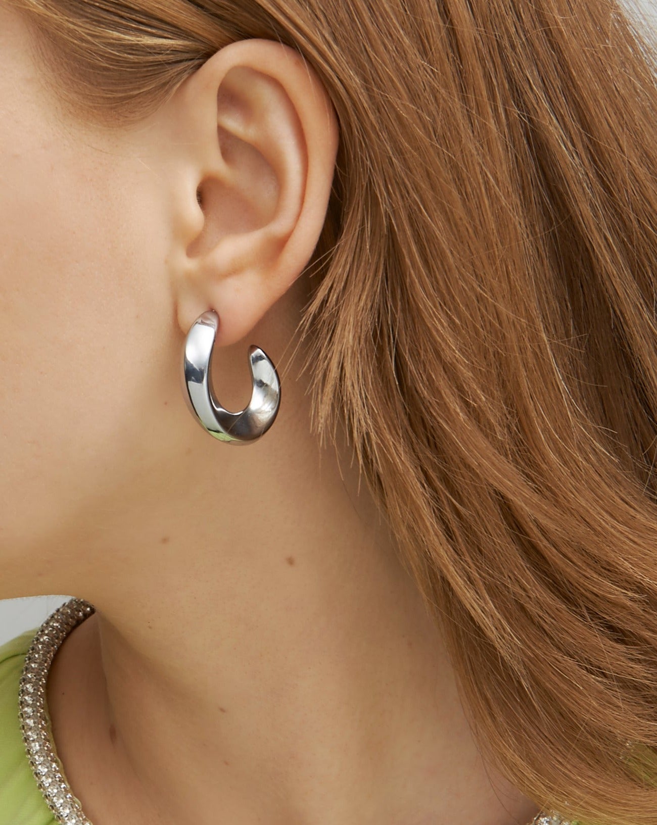 Close-up of a person with light brown hair wearing For Art's Sake® Wave Earrings Silver. The chunky half hoop design is thick and slightly curved, and the person sports a light green top with a silver collar. The focus is on the ear, earring, and a portion of the hair and clothing.