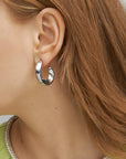 Close-up of a person with light brown hair wearing For Art's Sake® Wave Earrings Silver. The chunky half hoop design is thick and slightly curved, and the person sports a light green top with a silver collar. The focus is on the ear, earring, and a portion of the hair and clothing.