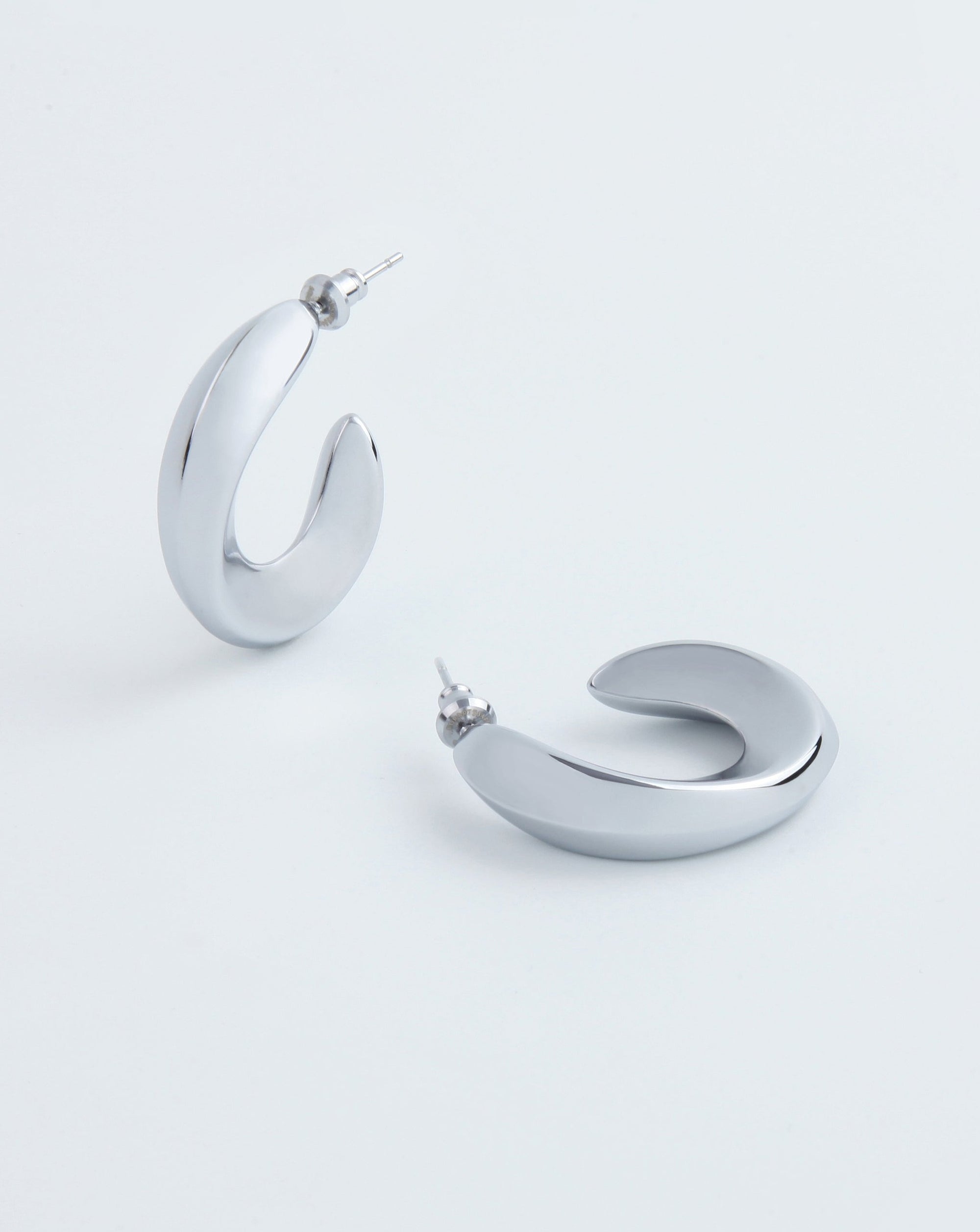 A pair of stylish, chunky Wave Earrings Silver by For Art's Sake® with a unique, open-ended design. The metallic surface has a polished, reflective finish. The earrings are positioned on a plain white background, one standing upright and the other lying flat.